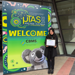 Linda Tian posing with Oral Presentation Award in front of MicroTAS 2025 banner