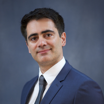 Professional headshot of Alireza Khaligh with short dark hair wearing a navy suit, white dress shirt and dark tie, smiling slightly against a neutral blue-grey background.